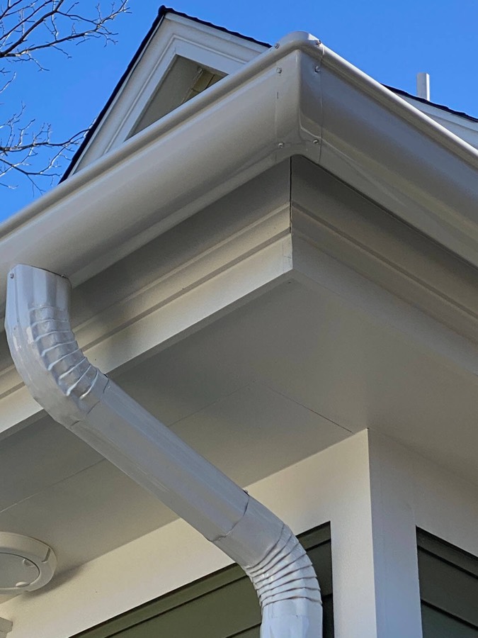 White gutter and downspout at the corner of a home's eave, directing roof runoff away from the fascia